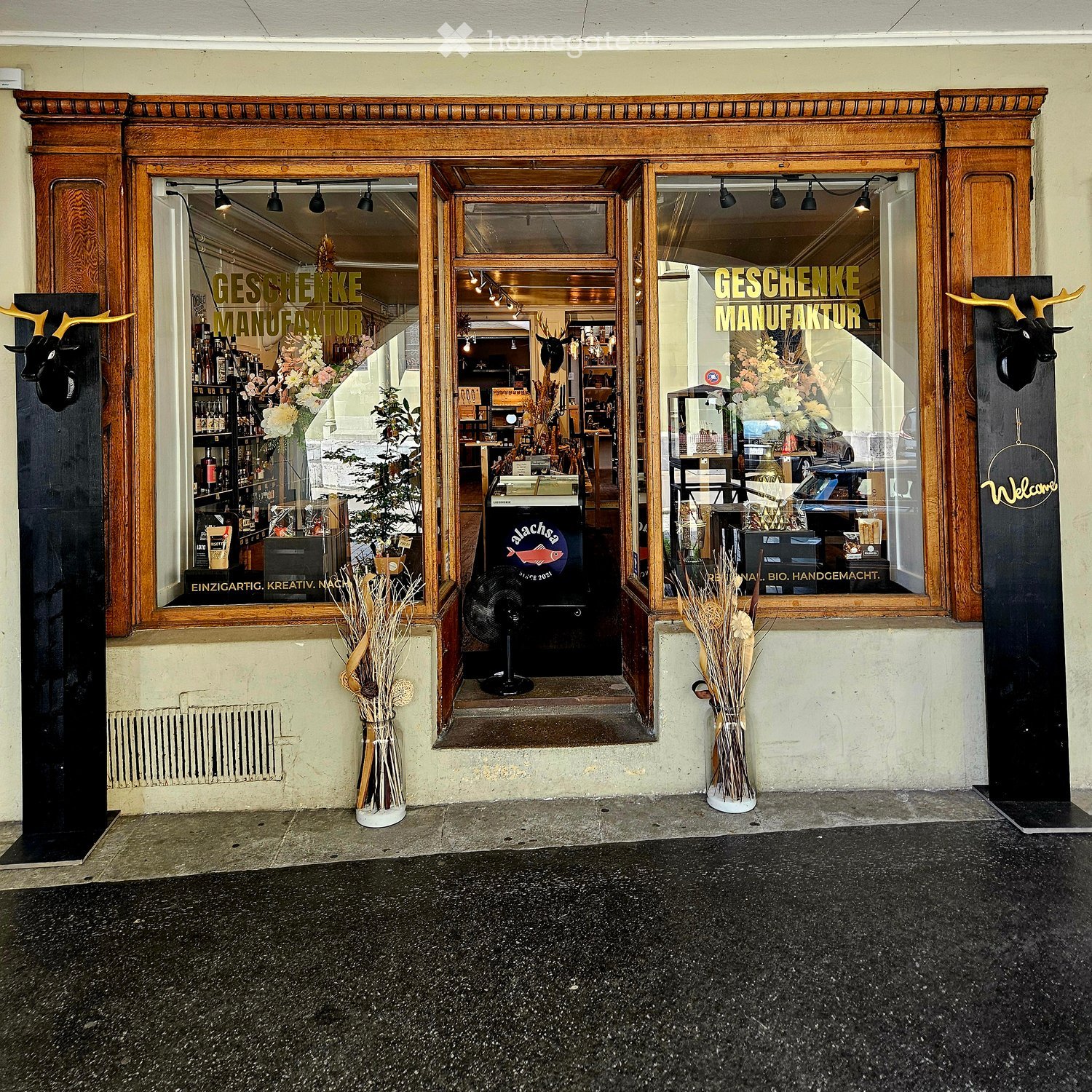 Storefront with large wooden-framed windows and doors, displaying the sign "GESCHENKE MANUFAKTUR" and interior shelves with various items, including bottles and decorative objects. The entrance has a small step and a fan placed near the door.