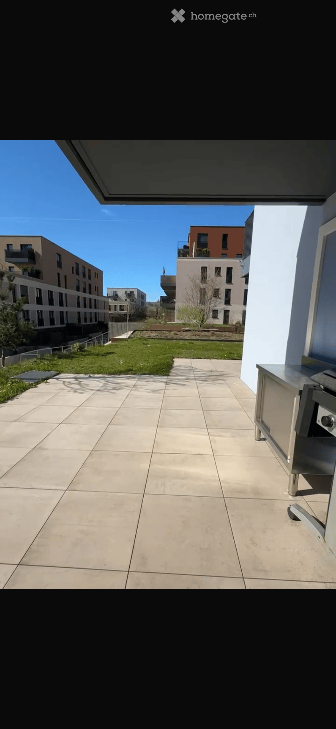 Spacious balcony with tiled floor, modern stainless steel railing, and a view of a landscaped area with grass and modern apartment buildings.