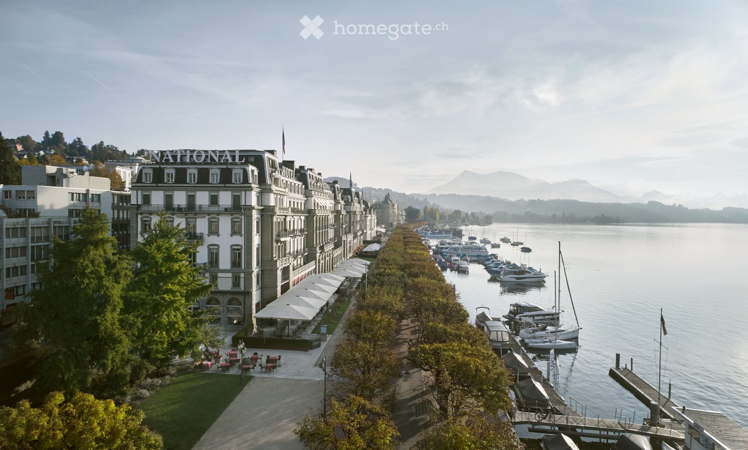 A large, elegant hotel named "National" is situated along a waterfront promenade, with numerous boats docked in the calm water. The building features classic architecture with multiple windows and balconies, and the surrounding area includes outdoor seati