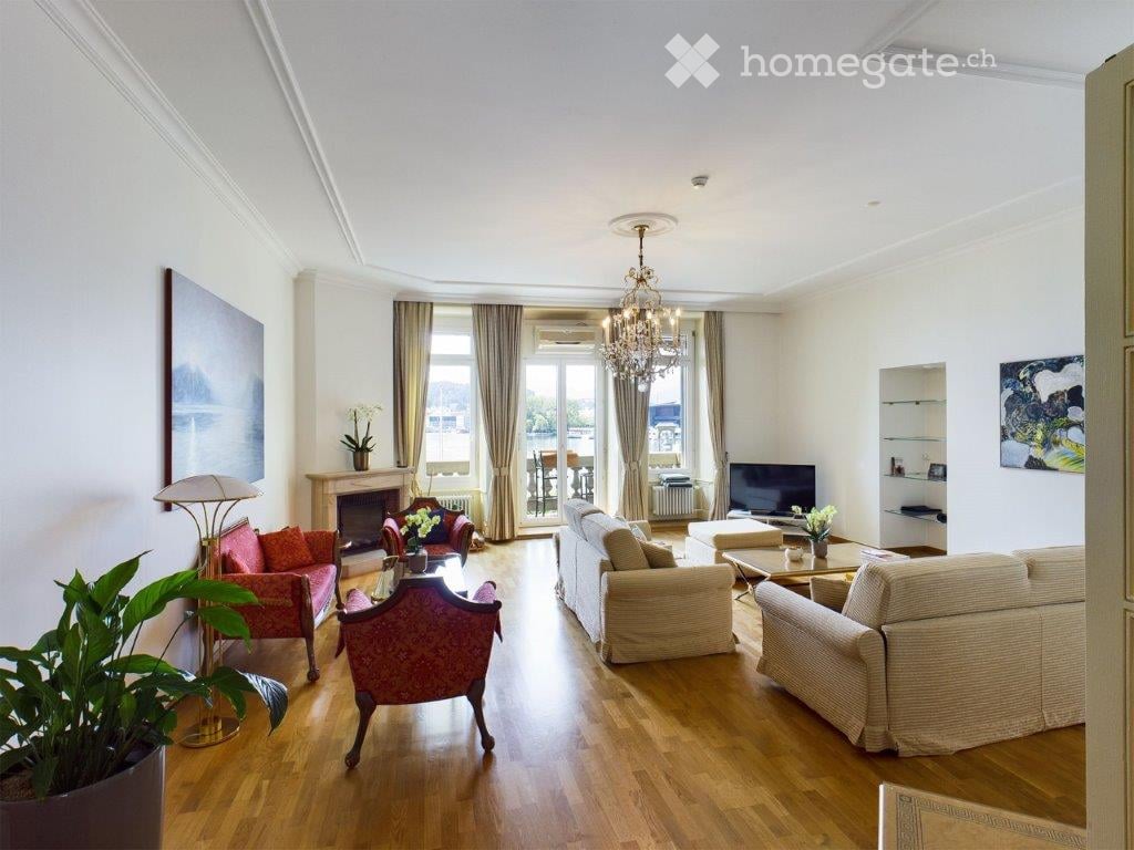 Elegant living room with wooden flooring, large windows offering a view, a fireplace, a chandelier, a sofa, armchairs, coffee table, and decorative plants.