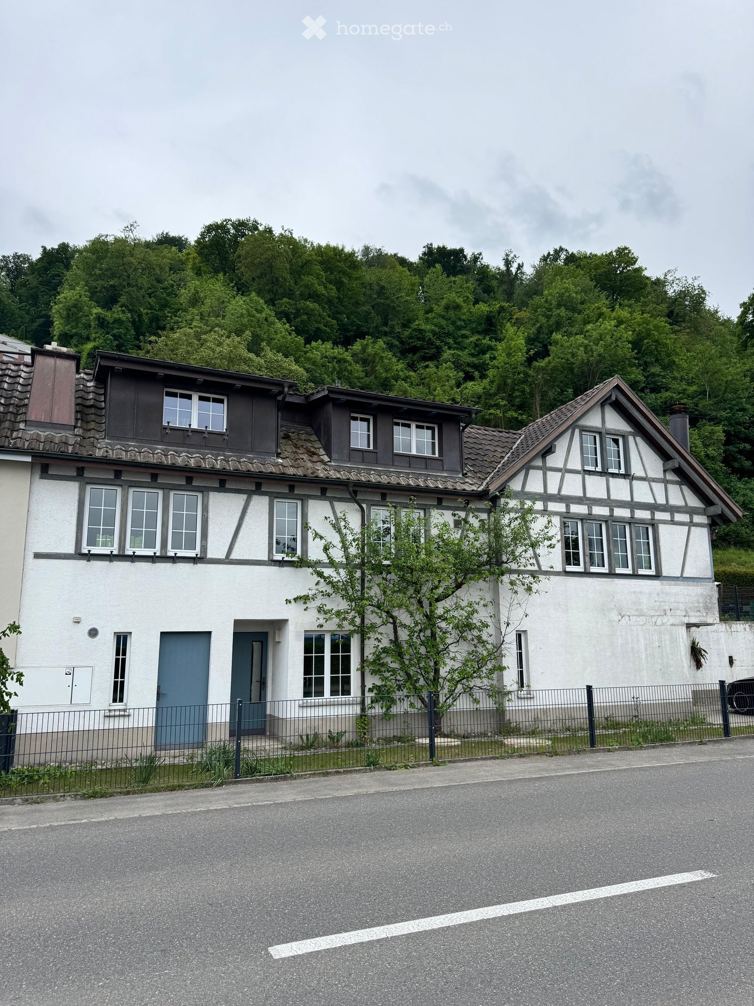 Two-story house with white walls, dark wooden trim, multiple windows, and a small tree in front.