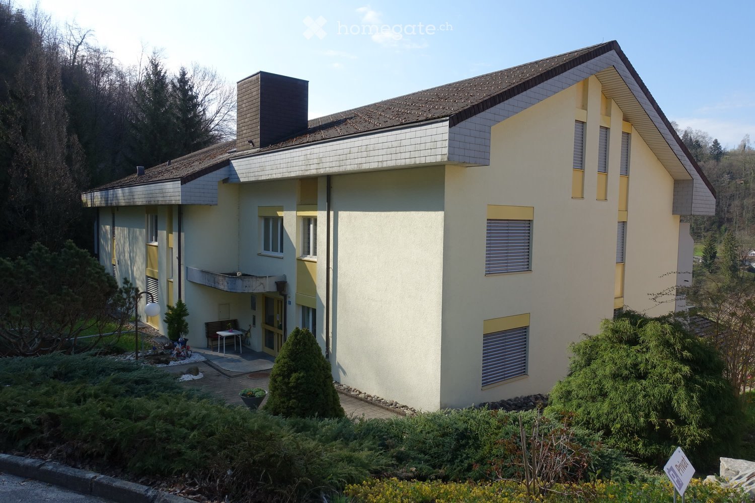 Two story house, white and yellow walls, brown roof, chimney, windows with shutters, plants and shrubs in front