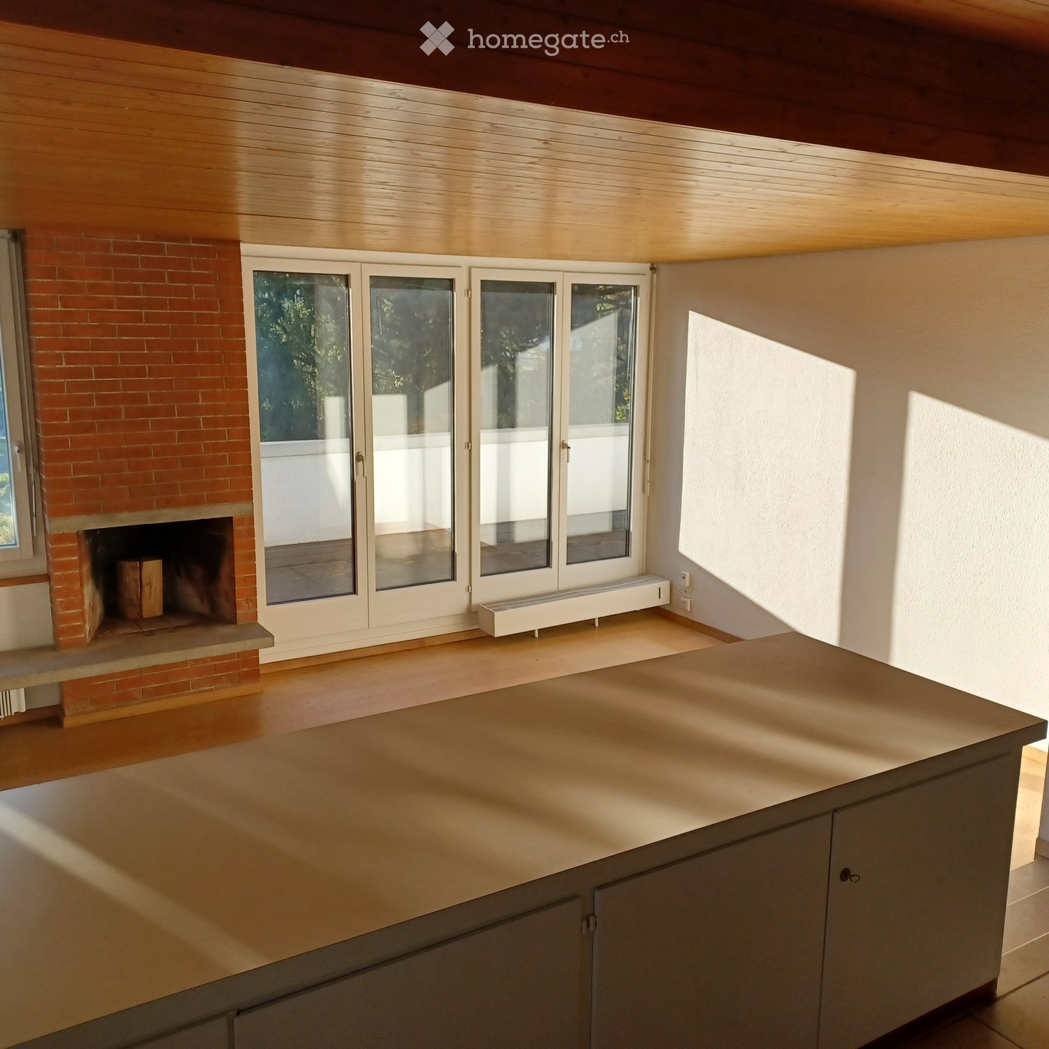 Bright, open room with wooden ceiling and floor, fireplace with brick surround, large glass doors leading outside, and a central table.