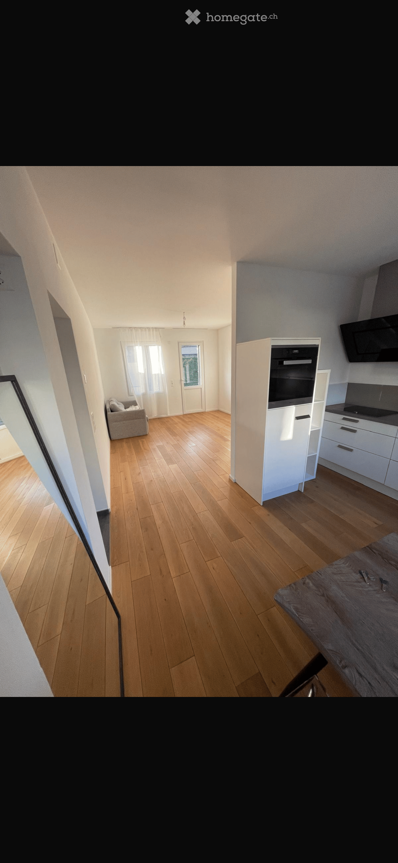 A modern, open-plan living and dining area with light wooden flooring, white walls, and large windows. The space includes a sofa, a kitchen area with cabinets and appliances, and a dining table.