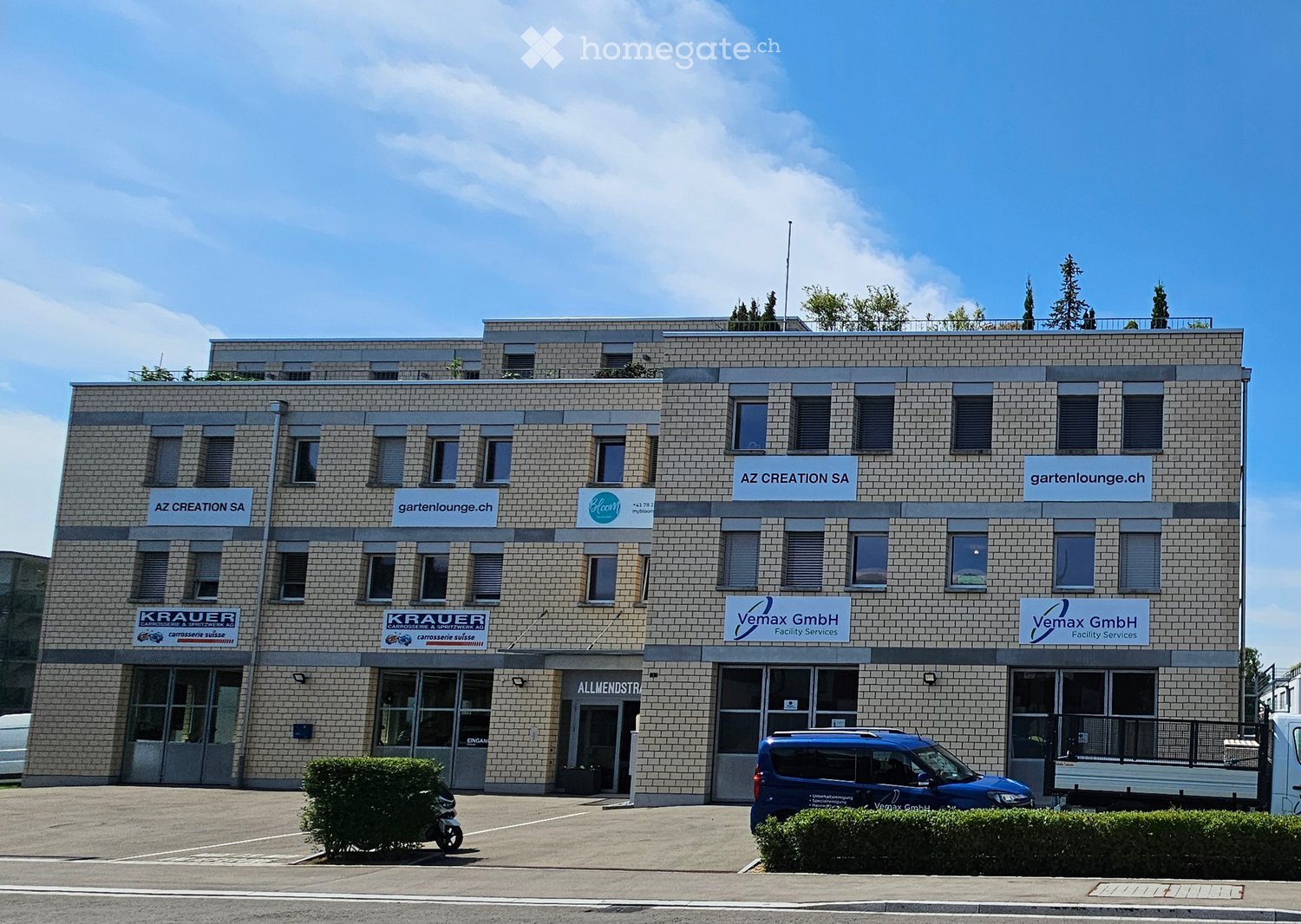 Building. A modern, multi-story commercial building with a brick facade, multiple company logos, and several windows. The building has a flat roof with greenery and is surrounded by a paved area with parked vehicles.