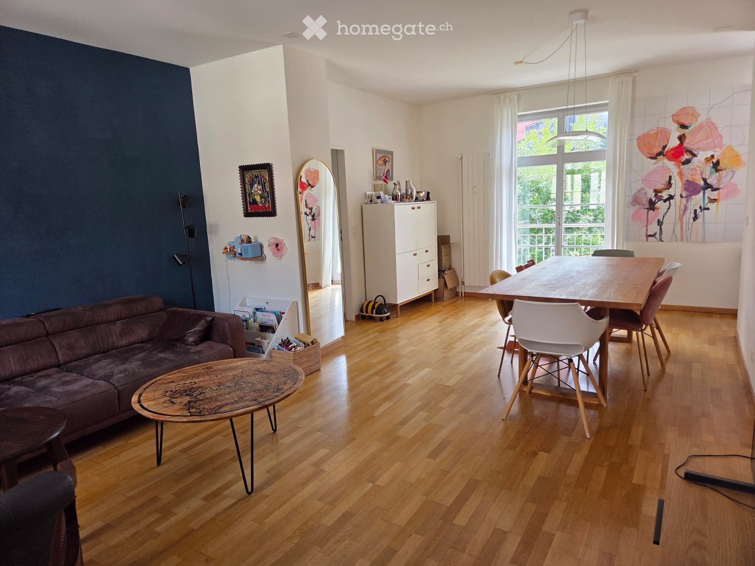 Open-plan living and dining area with wooden flooring, a dark blue accent wall, a brown leather sofa, a round wooden coffee table, a dining table with chairs, a white cabinet, and artwork on the walls.