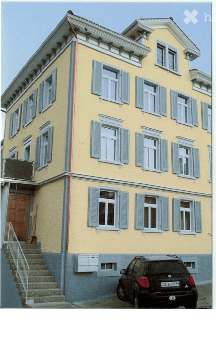 A three-story yellow building with multiple windows and shutters, a staircase leading to the entrance, and a parked car in front.