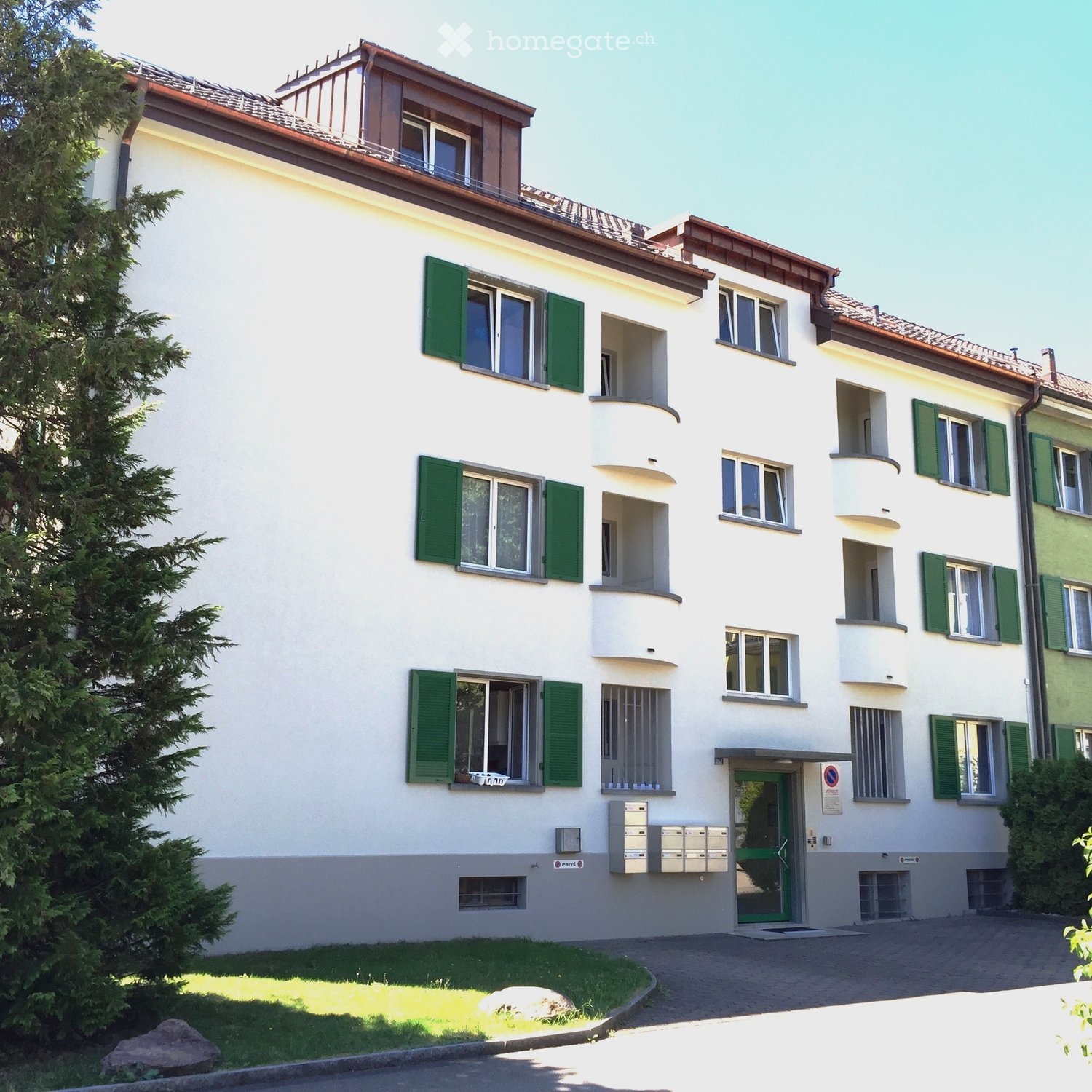 A three-story residential building with white walls, green shutters, and multiple windows. Features include balconies, a front entrance with a green door, and a small garden area with a tree and rock.