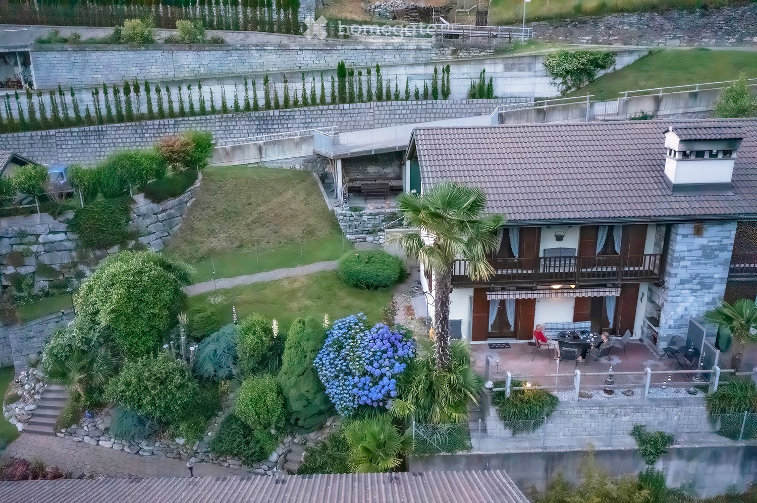 A two-story house with a tiled roof, surrounded by a well-maintained garden with various plants, trees, and a small patio area with outdoor furniture.