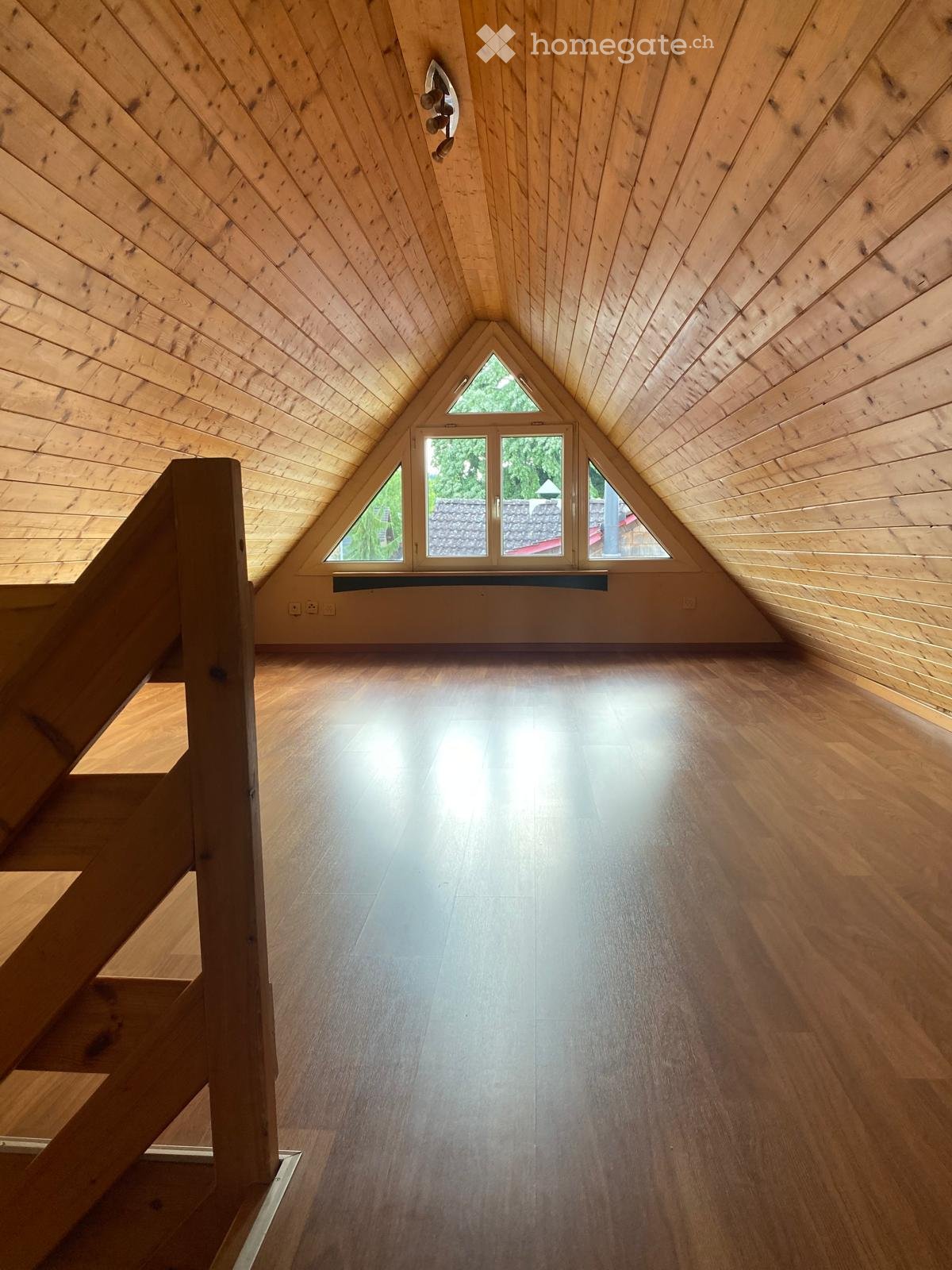 Bright, empty attic space with wooden walls, ceiling, and floor, featuring large triangular windows and a staircase leading up.