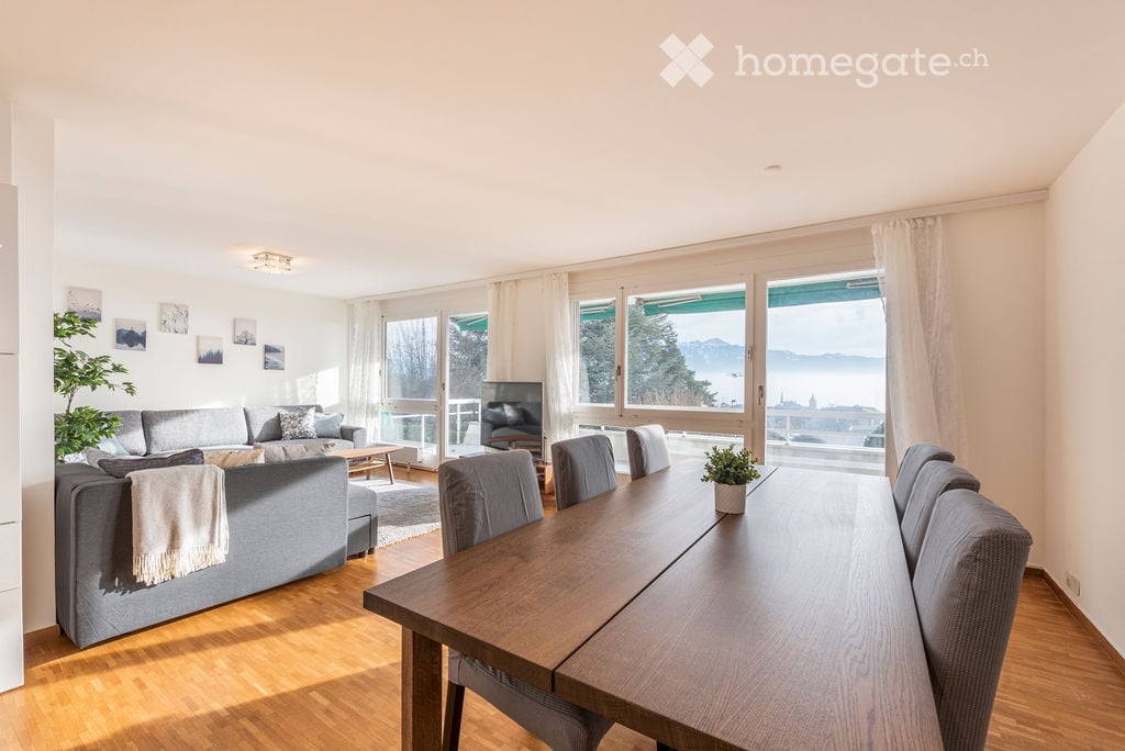 Furnished Room. Bright living and dining area with large windows offering a scenic view, featuring a grey sofa, coffee table, dining table with chairs, and a television.