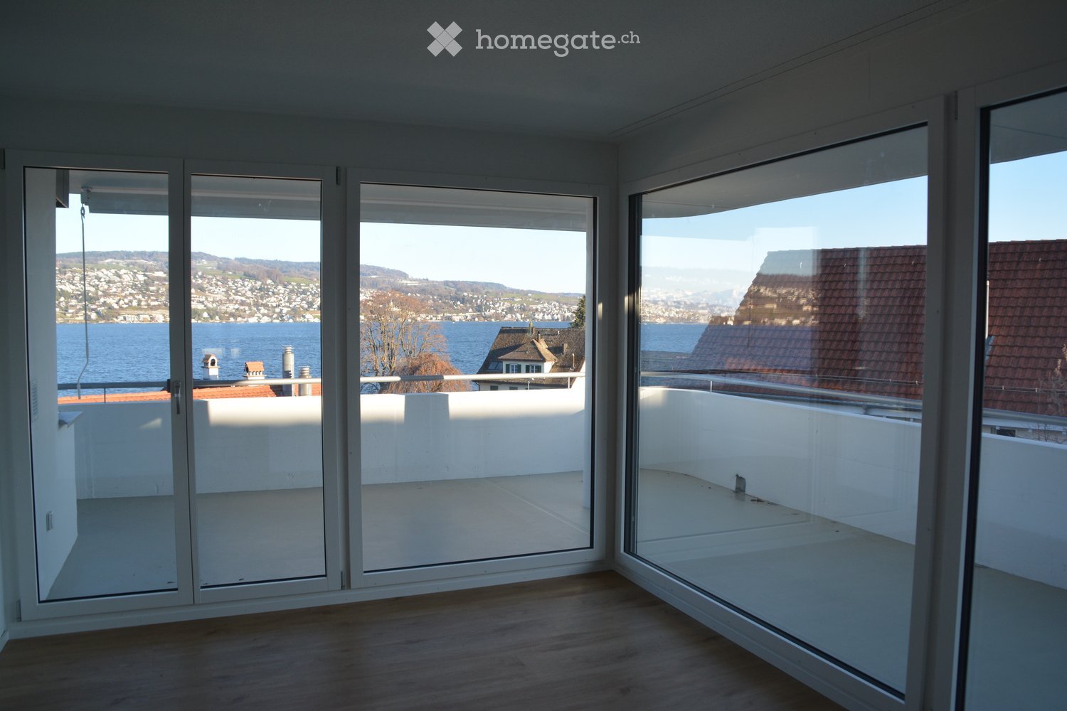 Empty room with large glass doors opening to a balcony, overlooking a scenic lake and distant hills.