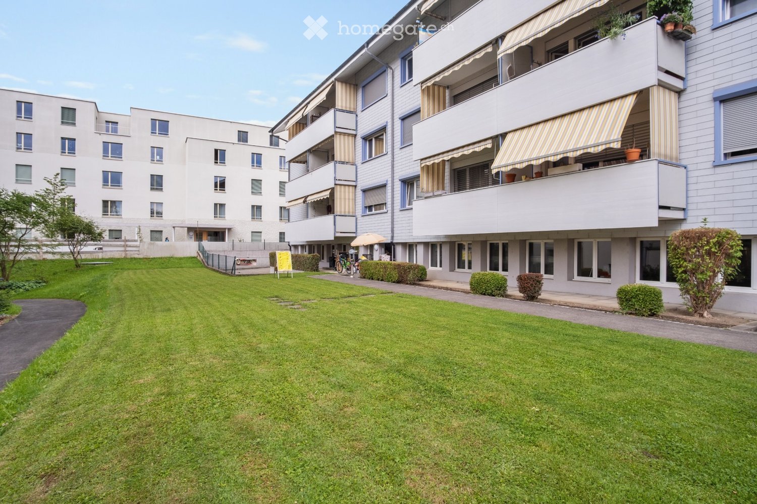 Multi-story residential building with balconies, striped awnings, and a well-maintained lawn in front.