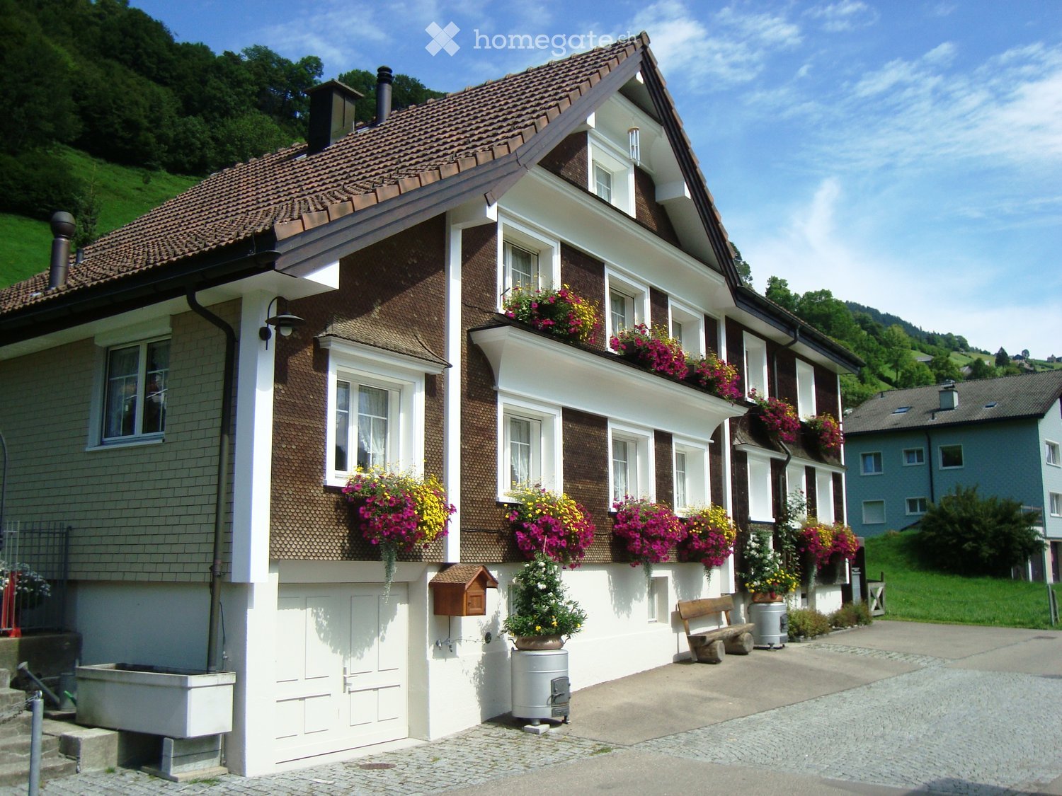 Two-story house with brown tiled roof, white trim, multiple windows adorned with colorful flower boxes, and a small garden area with a bench and potted plants.