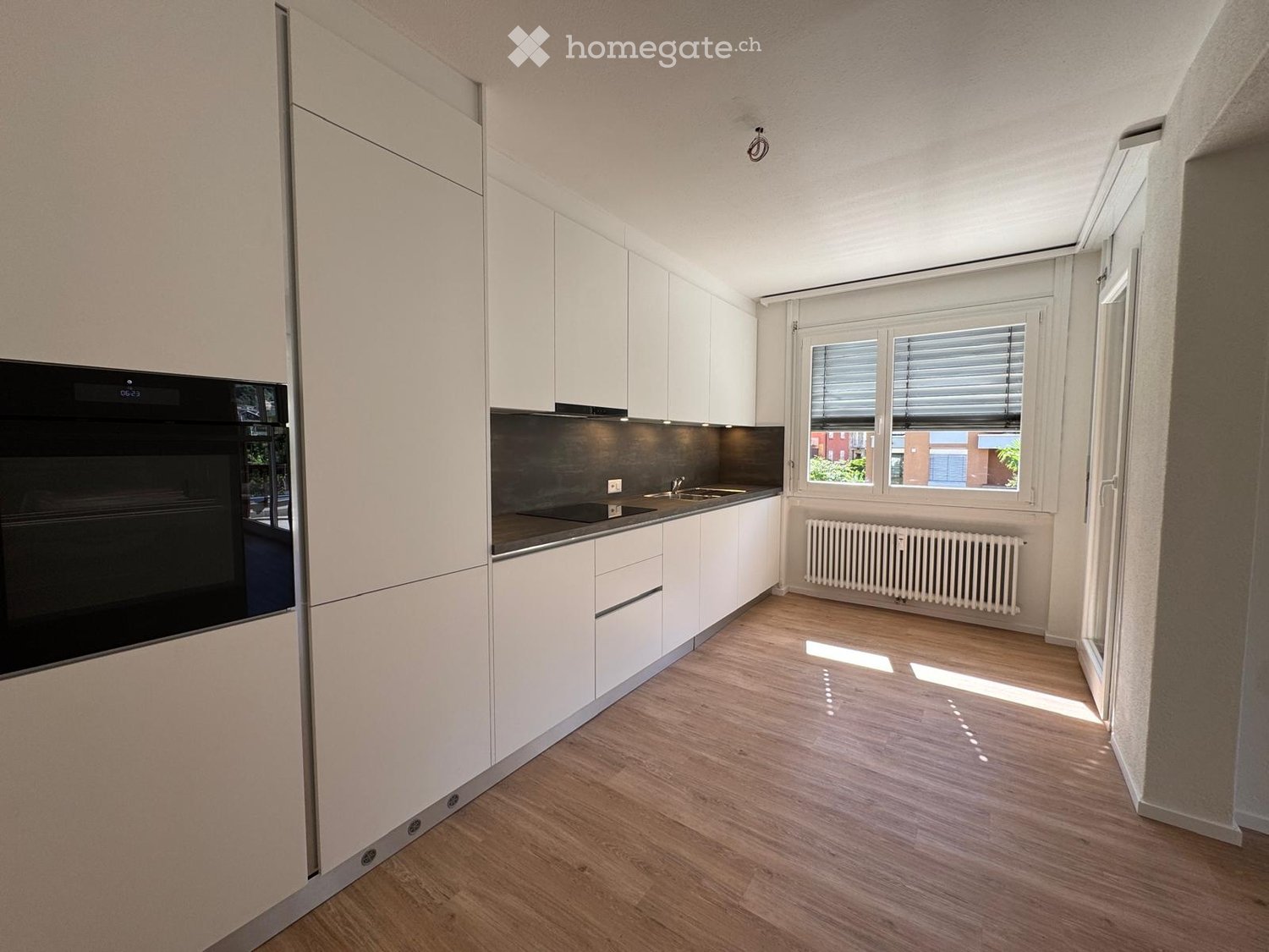 Modern kitchen with white cabinets, wooden flooring, and a black oven. Features a sink, radiator, and large windows with blinds.