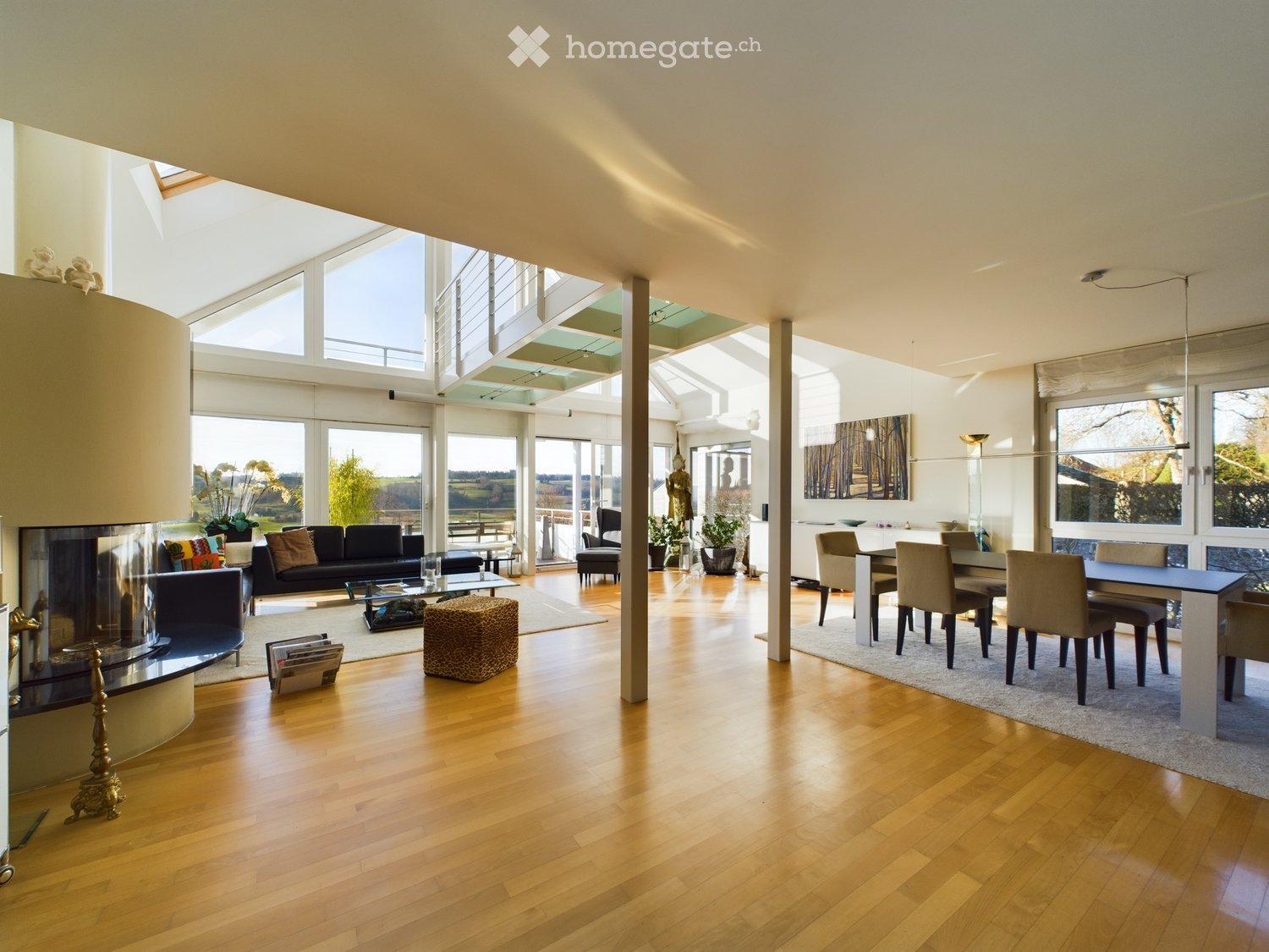 Open-plan living and dining area with wooden flooring, large windows offering a scenic view, a fireplace, a sofa, a coffee table, a dining table with chairs, and decorative elements.