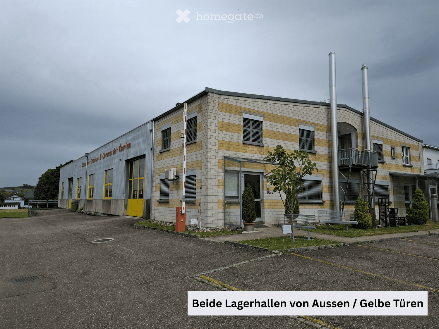 Two-story industrial building with yellow doors, tiled facade, and ventilation pipes. Small trees and benches are visible in front.