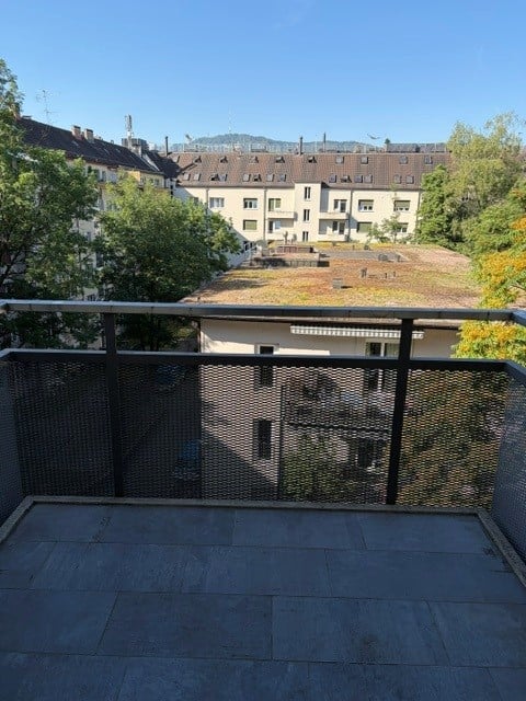 Balcony with metal railing, overlooking a cityscape with residential buildings and greenery.