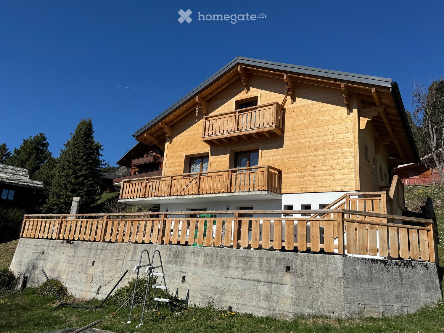 A two-story wooden house with balconies, situated on a raised ground floor with a concrete retaining wall.