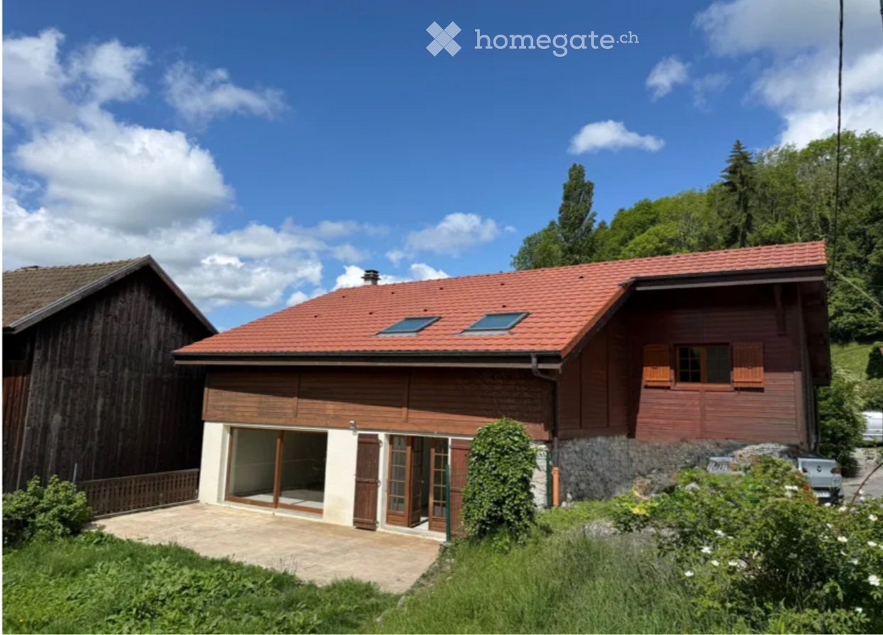 Two-story wooden house with red-tiled roof, large windows, and a covered entrance area. The house is surrounded by greenery and has a stone foundation.