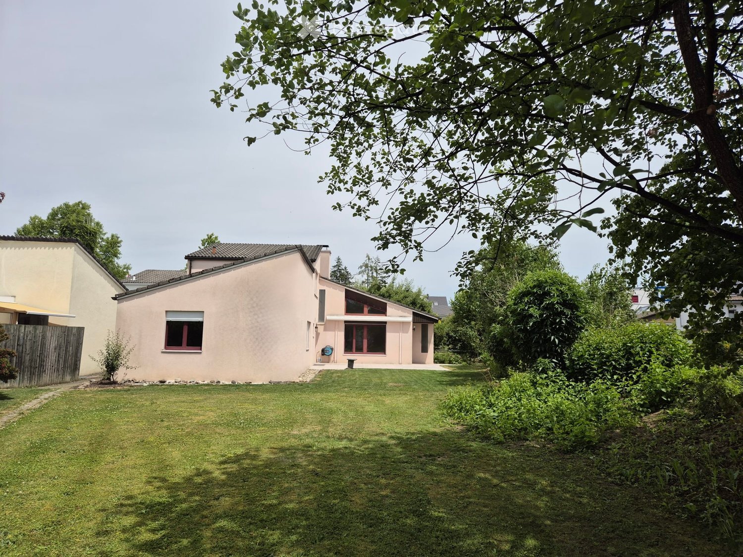 A well-maintained lawn in front of a modern house with a pink facade, large windows, and a wooden fence on the left side.