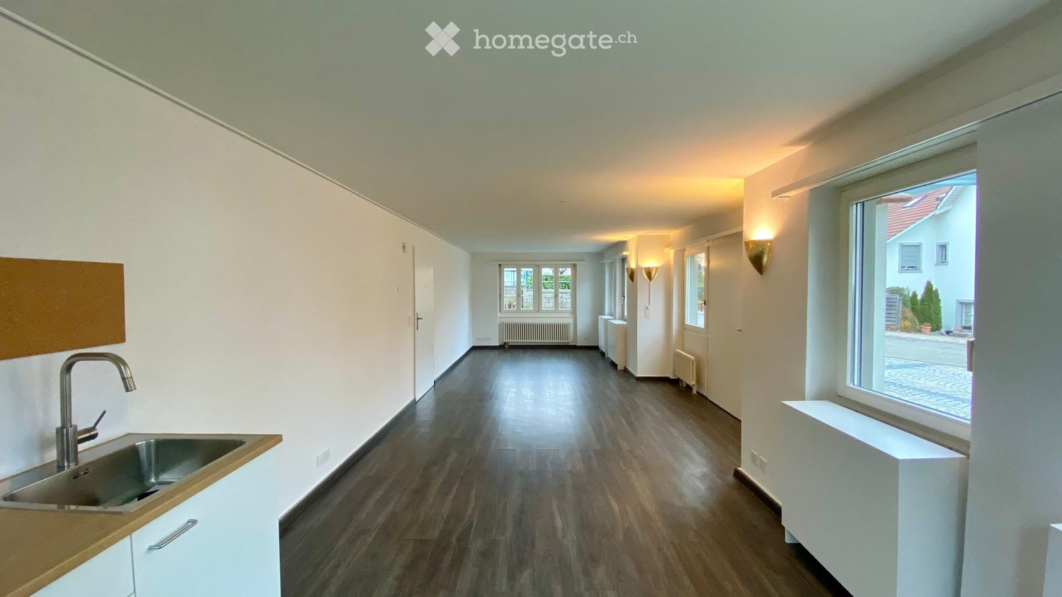 Long narrow room with wooden floors, white walls, and large windows. Sink on left, radiators on right.