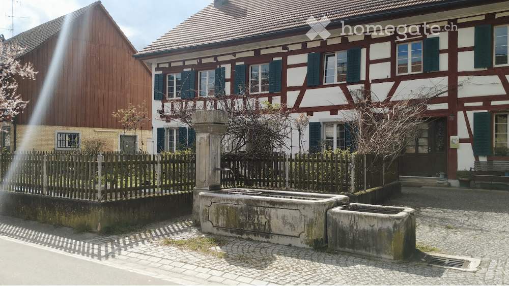 Traditional half-timbered house with white and red wooden framing, multiple windows with shutters, and a tiled roof. A stone well and wooden fence are visible in the foreground.