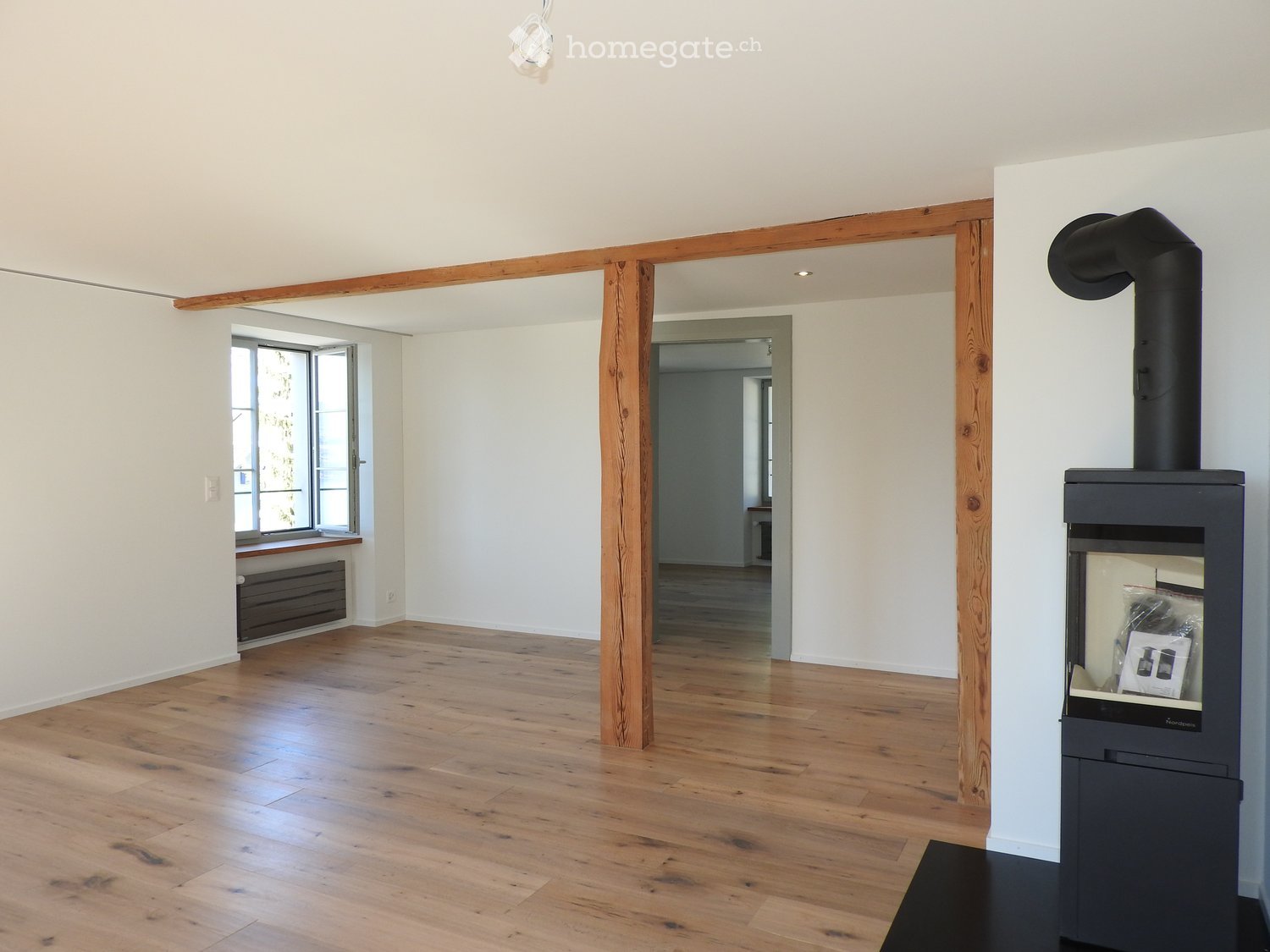 Empty room with wooden flooring, white walls, and a modern black wood-burning stove with a chimney. The room features exposed wooden beams and a large window with a view.