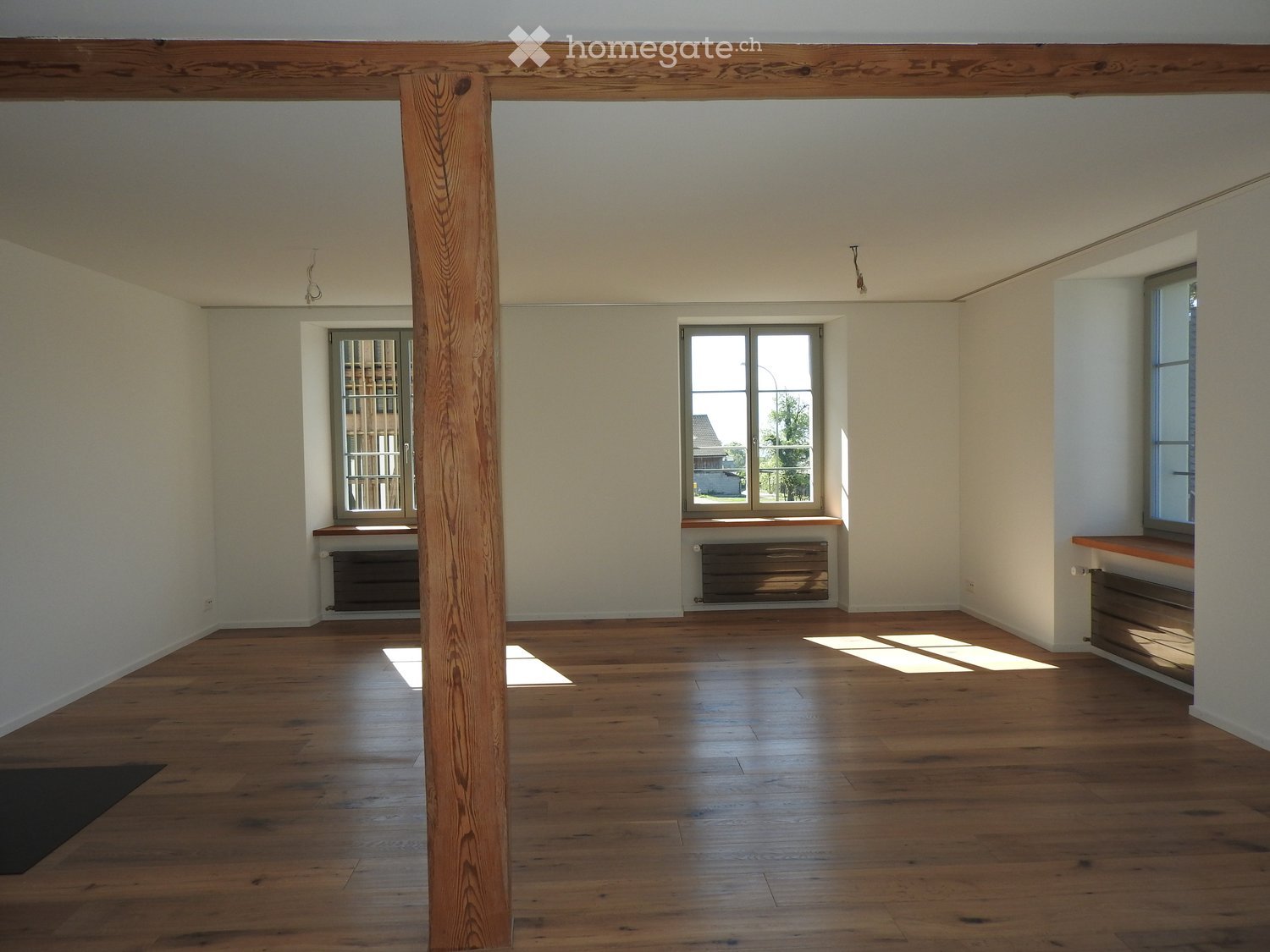 Empty room with wooden flooring, white walls, and large windows allowing natural light. Features a central wooden pillar and radiators beneath the windows.