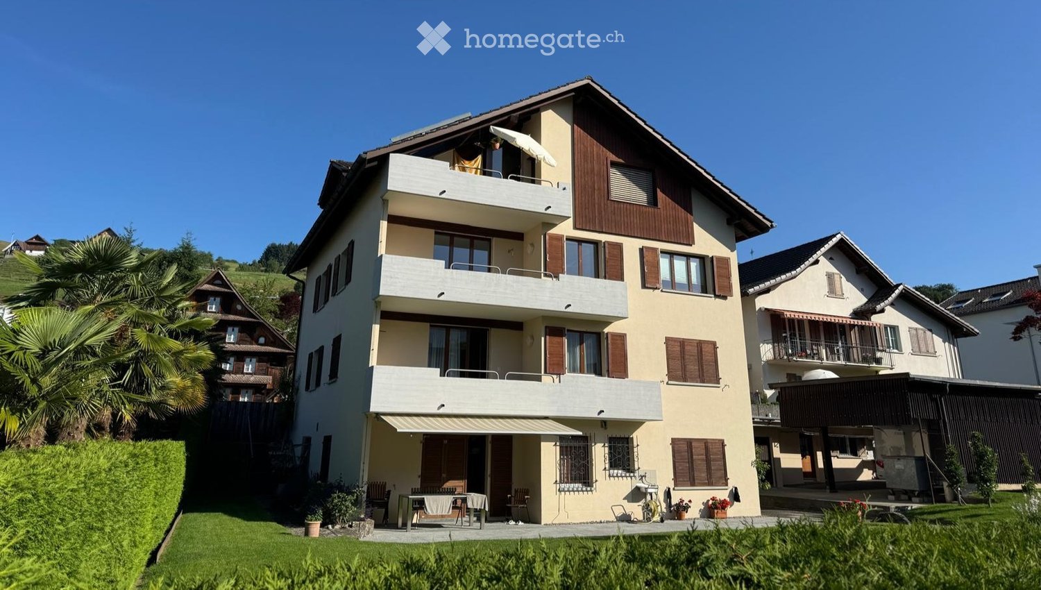 A two-story house with beige walls, wooden shutters, and multiple balconies. The house features a well-maintained garden with a patio area, outdoor furniture, and potted plants.