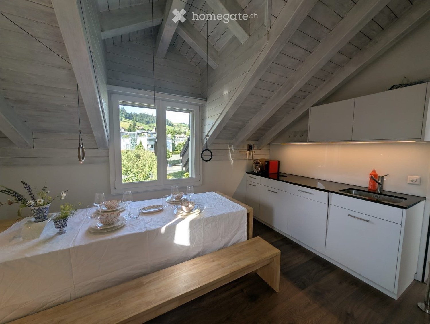 A modern, open-plan kitchen and dining area with white cabinets, a black countertop, a wooden dining table with a bench, and a window overlooking a scenic landscape.
