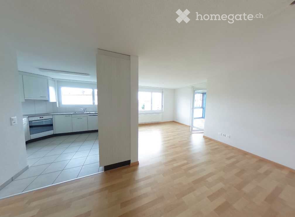 Empty room with wooden flooring and white walls, connected to a small kitchen area with white cabinets and appliances.