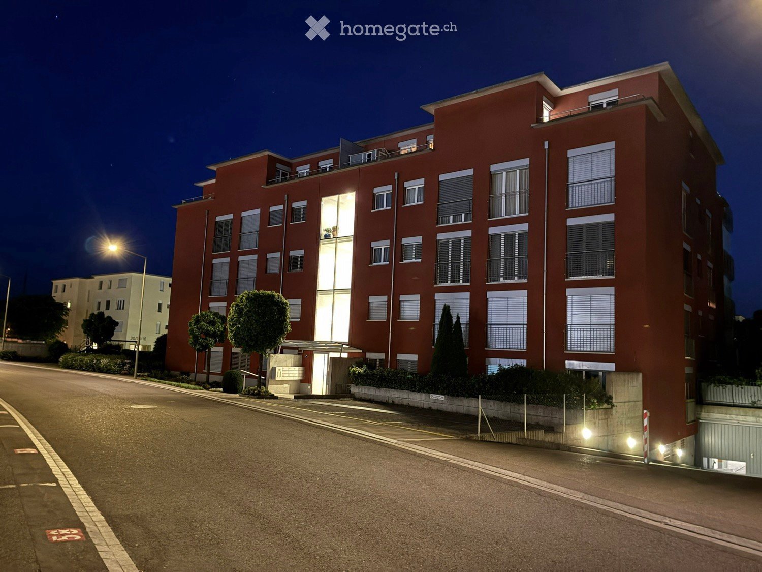 Four-story red apartment building with illuminated windows and balconies, surrounded by trees and a street.