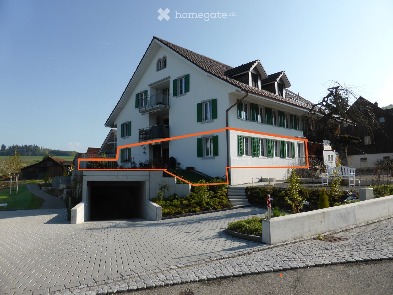 Two-story white house with green shutters, multiple windows, and a garage entrance. The house features a well-maintained garden and a paved driveway.