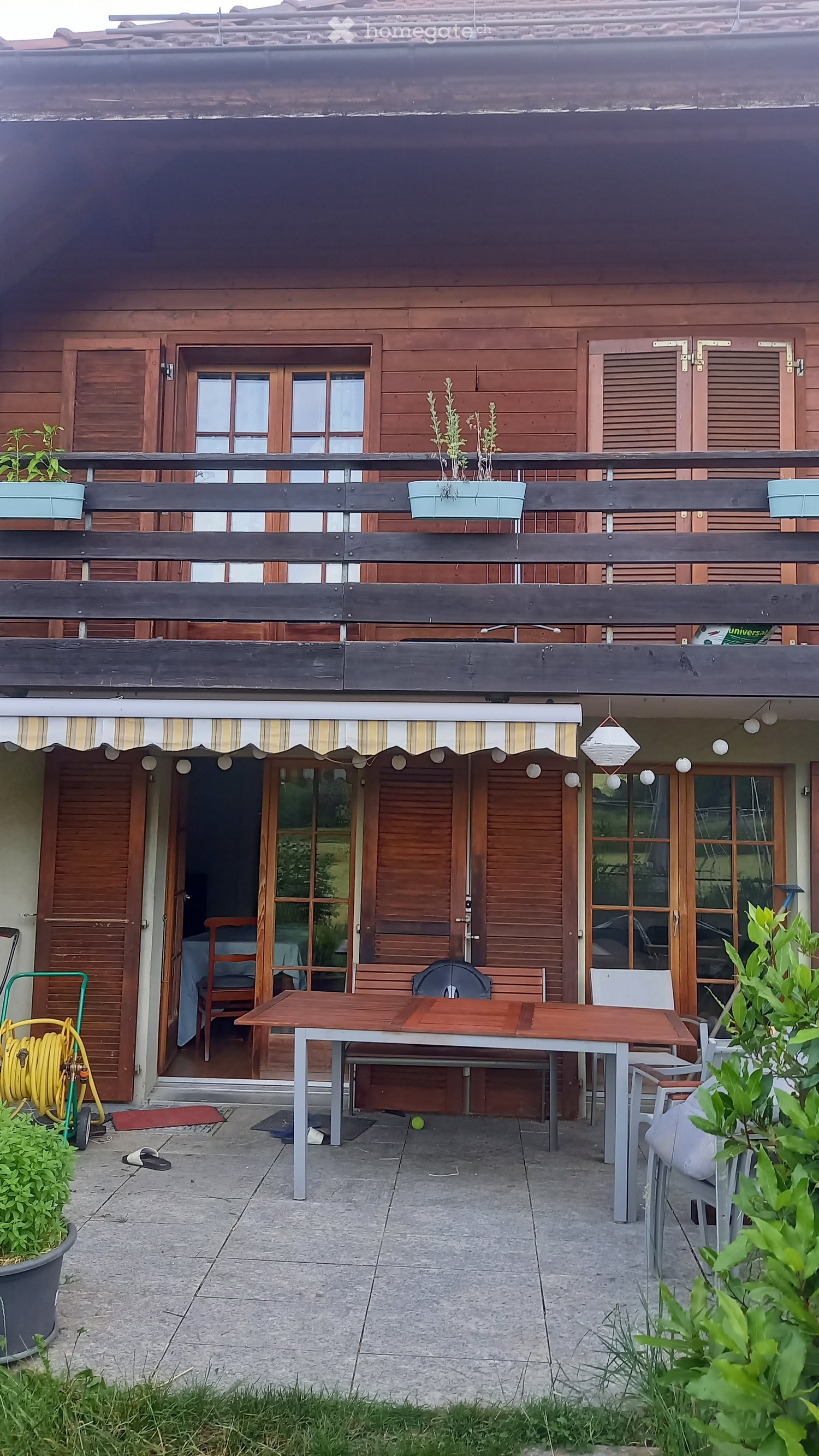 Balcony with wooden table, chairs, and bench, overlooking a wooden house with shutters and potted plants.