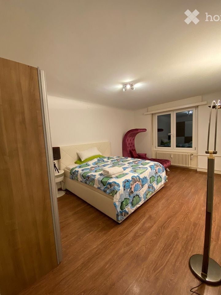 Cozy bedroom with wooden flooring, a large bed with a colorful duvet, a nightstand with a lamp, a red chair, and a radiator under the window.