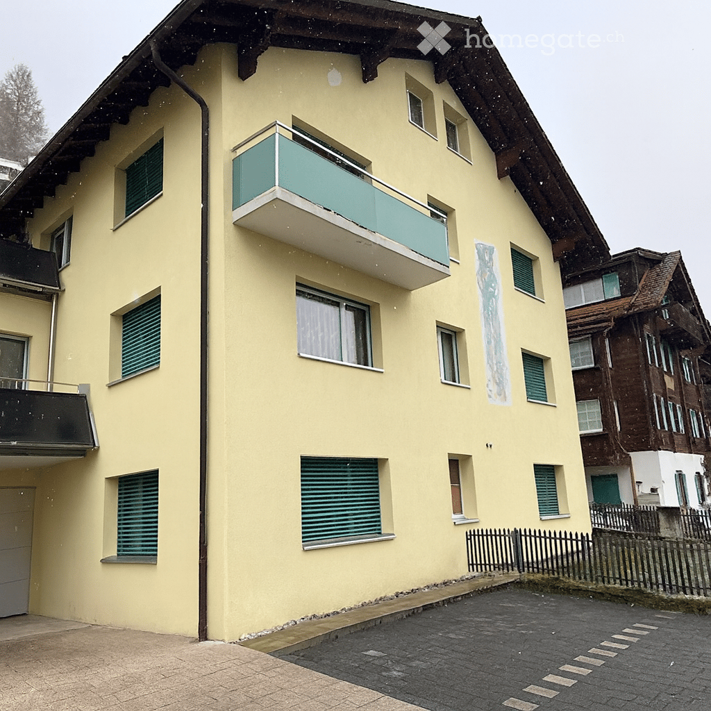 A building with yellow walls and green shutters, multiple windows, a small balcony on the front, and a roof entrance