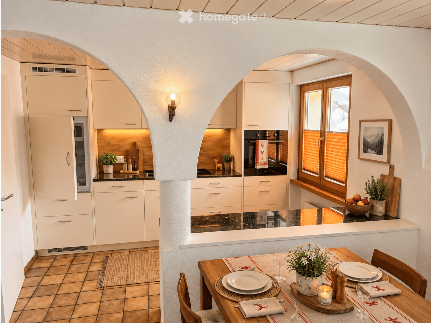Modern kitchen with white cabinets, a dining area, and a window view. The dining table is set with plates, wine glasses, and a potted plant. The kitchen features a microwave, oven, and refrigerator.
