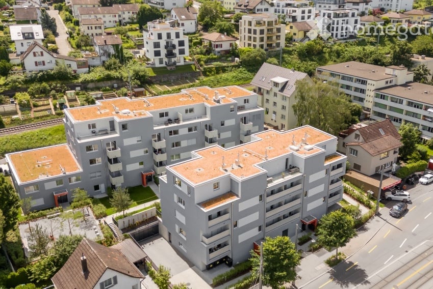 Aerial view of a modern multi-story residential building with grey facade and orange-tiled roofs, surrounded by other buildings and greenery.