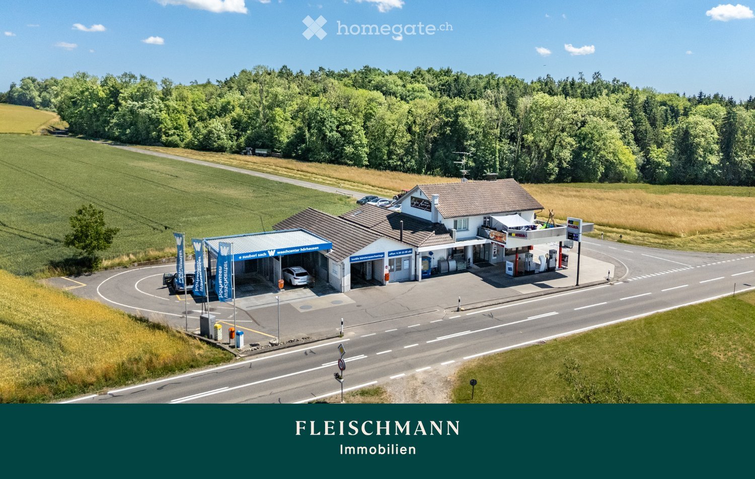 A gas station with a small building, multiple fuel pumps, and a covered area for vehicles. The station is situated near a road with surrounding fields and trees.