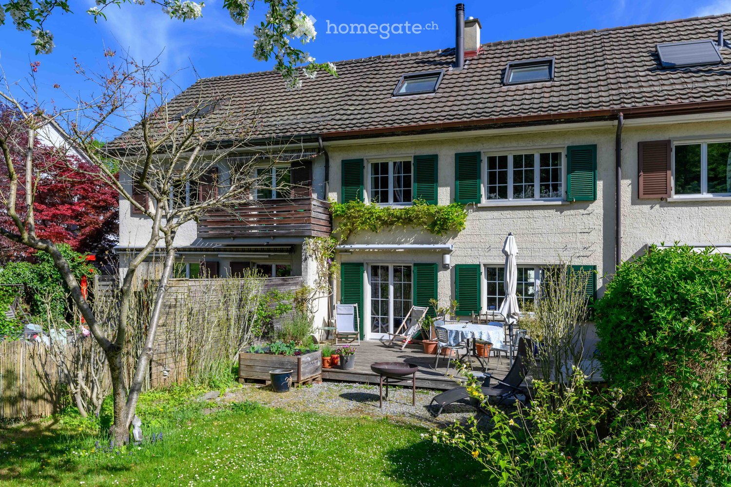 A two-story house with a well-maintained garden, featuring a patio area with outdoor furniture, potted plants, and a wooden fence.