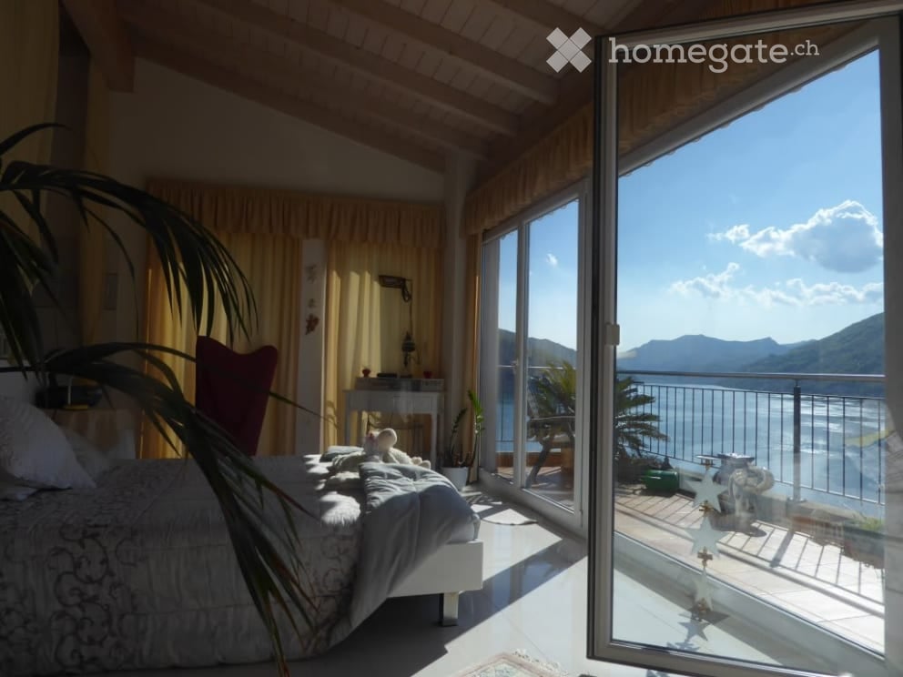 Cozy bedroom with large windows overlooking the lake and mountains, wooden ceiling, palm tree decor, and a comfortable-looking bed
