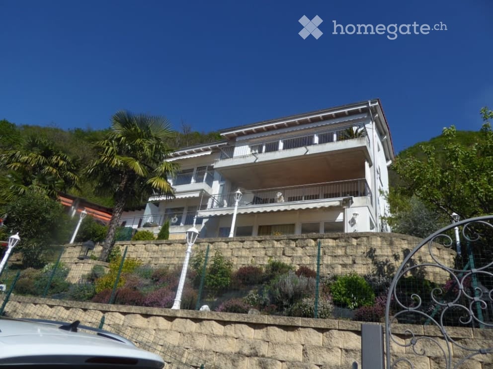 3-story apartment building with balconies, surrounded by palm trees and landscaping, with a stone retaining wall in the foreground