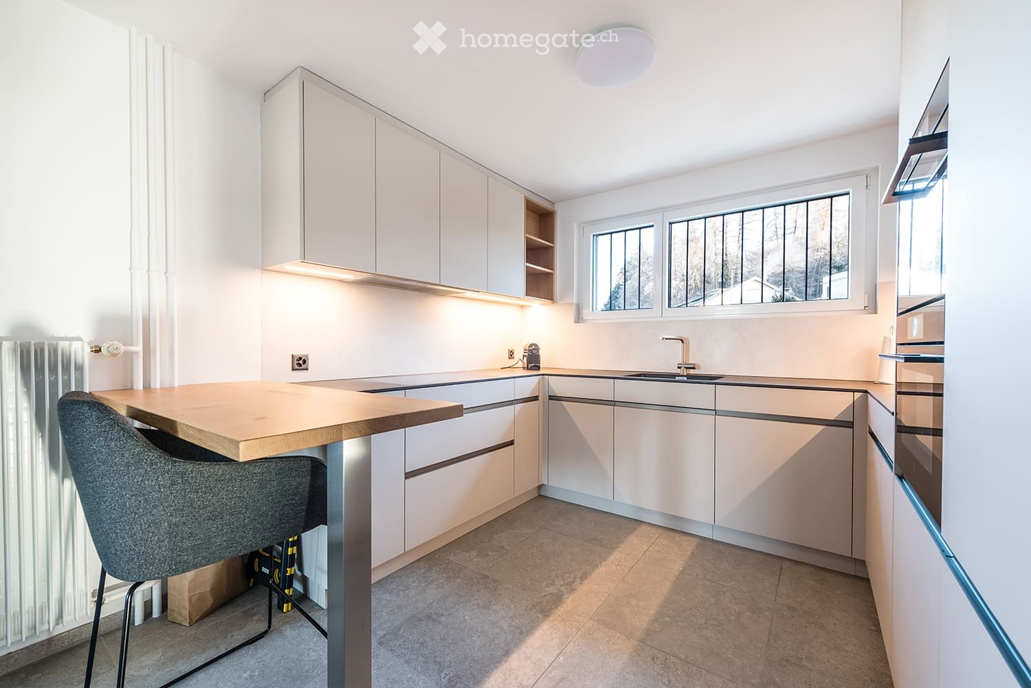 Modern kitchen with white cabinets, a wooden island, a sink, and a chair. The space features large windows and built-in lighting.