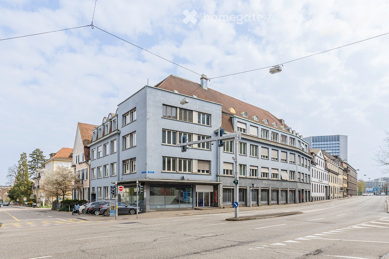 Multi-story building with light blue facade, multiple windows, and a curved corner section. The building is situated at a street corner with traffic lights and a few parked cars visible.
