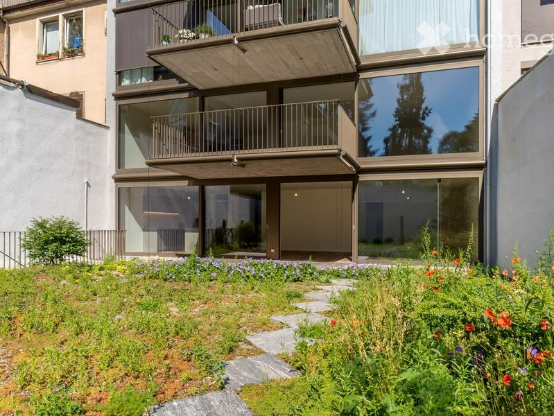Modern multi-story building with large glass windows and balconies, surrounded by a landscaped garden with a stone pathway and colorful flowers.