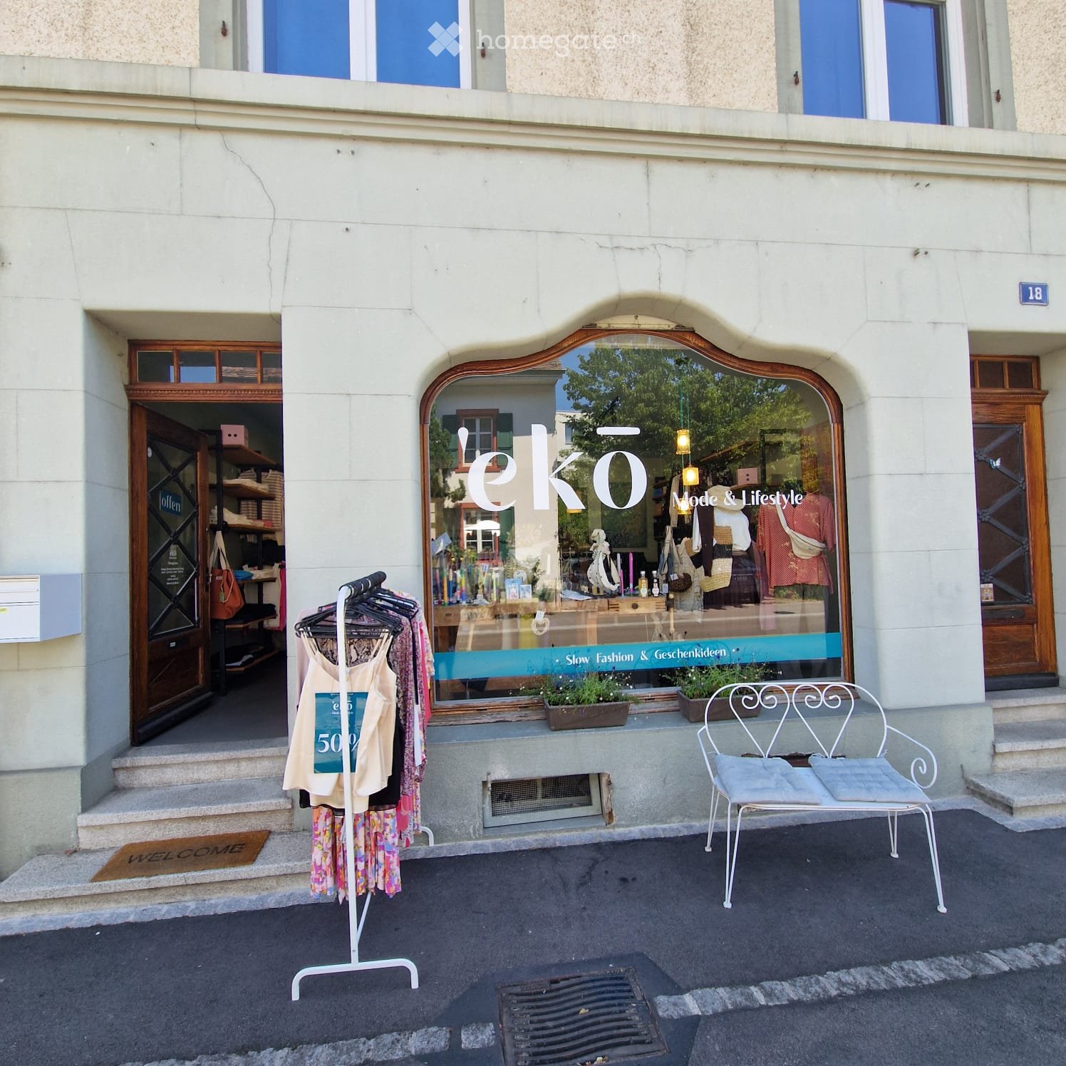 Facade of a two-story building with a storefront labeled "eko" featuring fashion and lifestyle items. Visible elements include a clothing rack with garments, a white metal chair, and a "WELCOME" doormat.