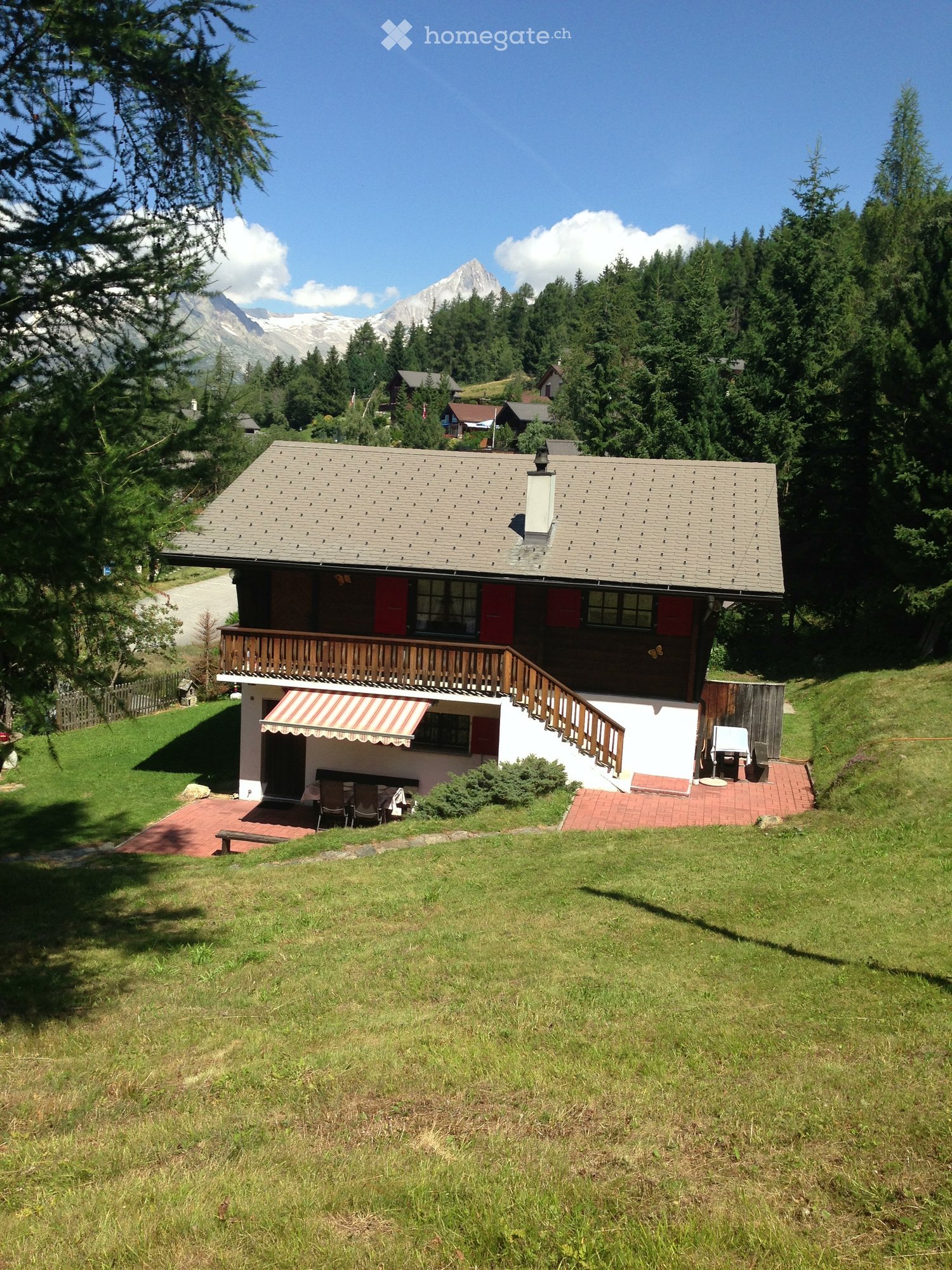 A two-story wooden house with a gray tiled roof, red shutters, and a balcony. The house is surrounded by a well-maintained lawn and features outdoor seating areas with tables and chairs.