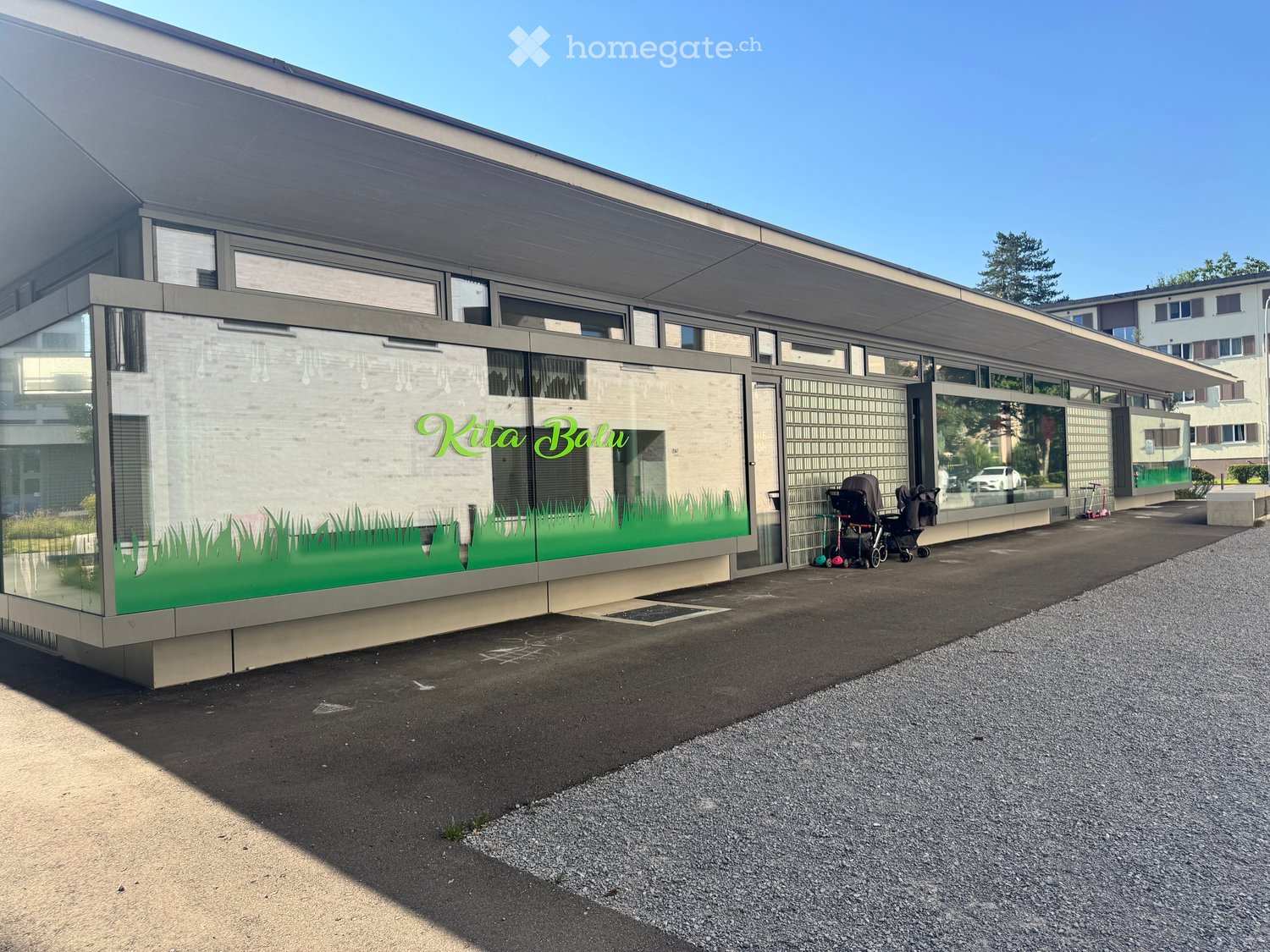 Modern building with large windows and a green mural featuring the text "Kita Balu" and a grassy design. The building has a flat roof and is surrounded by a paved area with a few strollers and scooters.