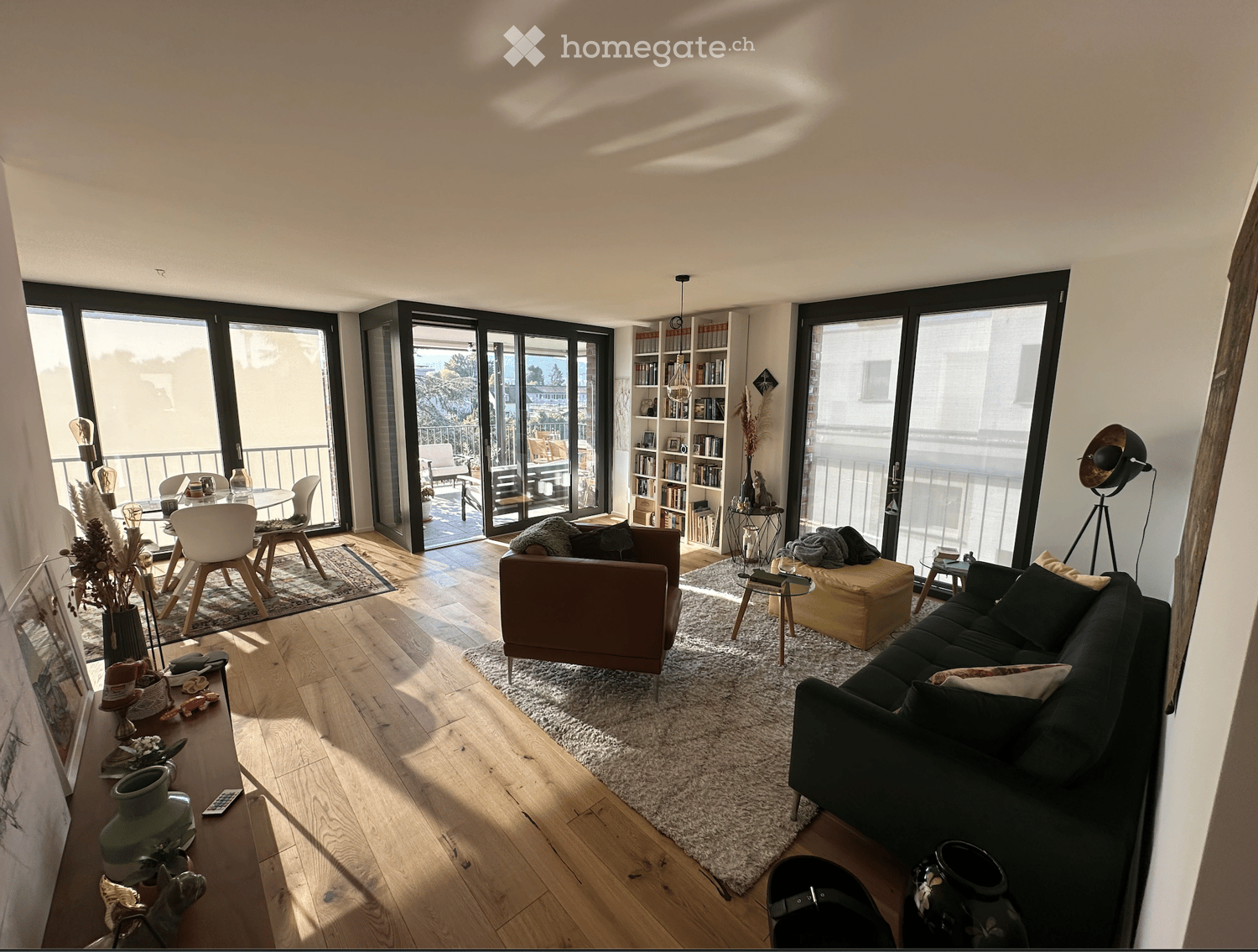 Modern living room with large windows and balcony access, featuring a dark grey sofa, brown armchair, coffee table, dining area with chairs, bookshelf, and decorative items.